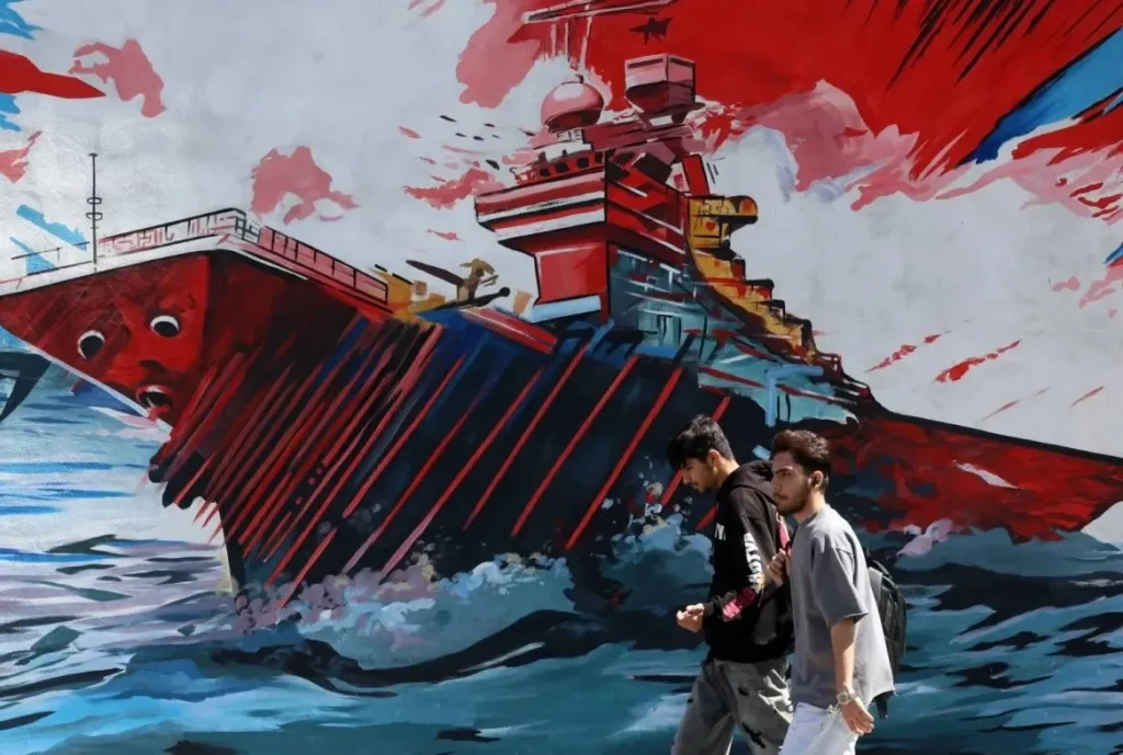رجلان يمران أمام لوحة جدارية مناهضة للولايات المتحدة وإسرائيل في طهران وتبين سفينة في مضيق هرمز (إ.ب.أ)