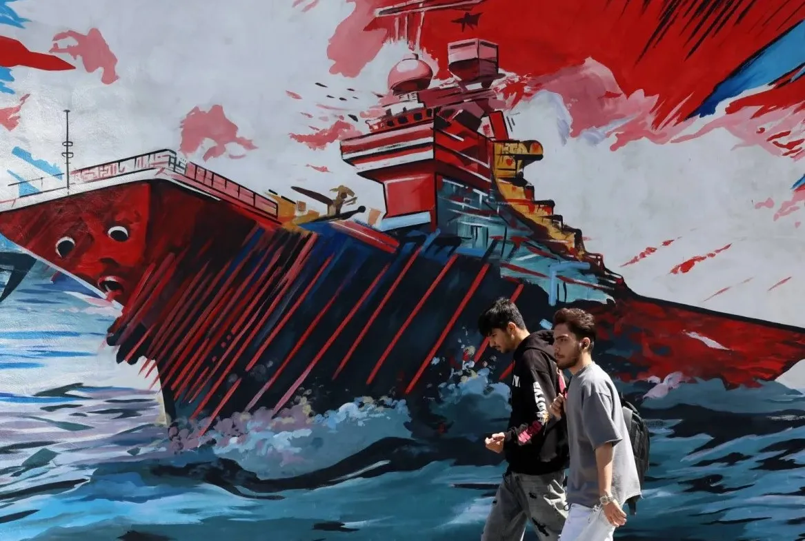 رجلان يمران أمام لوحة جدارية مناهضة للولايات المتحدة وإسرائيل في طهران وتبين سفينة في مضيق هرمز (إ.ب.أ)